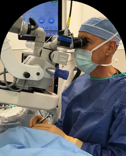Surgeon performing eye surgery using a microscope, wearing surgical attire and gloves.
