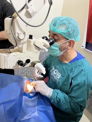 Doctor performing eye surgery using a microscope, wearing protective gear.